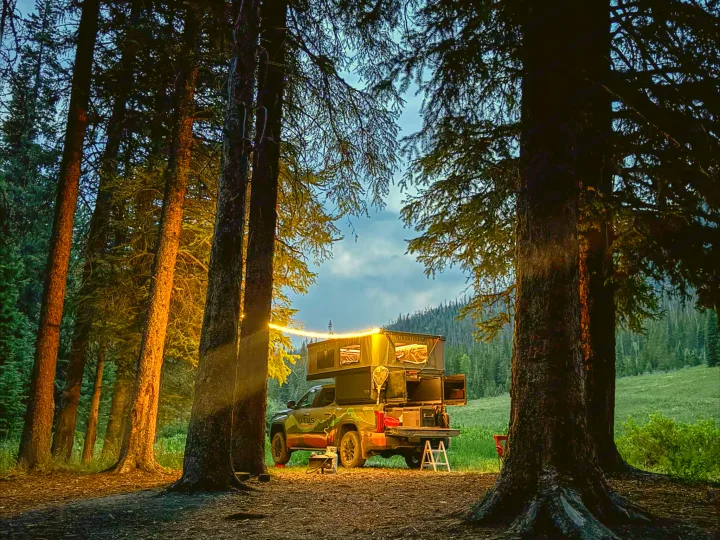 This illuminated truck camper RV rental is set up at a forest campsite, framed by tall trees under a twilight sky.. Toyota Tundra 2026