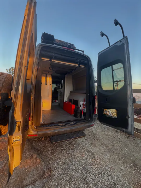 Adventure-ready RV rental: grey camper van with open rear, revealing custom interior, bed, storage, tools, & step.. Winnebago Revel 2022