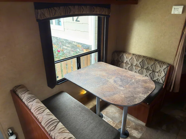 Inside an RV rental: a cozy dinette with a speckled table, striped benches, and a window view of a house and garden.. Jayco Jay Flight 2013