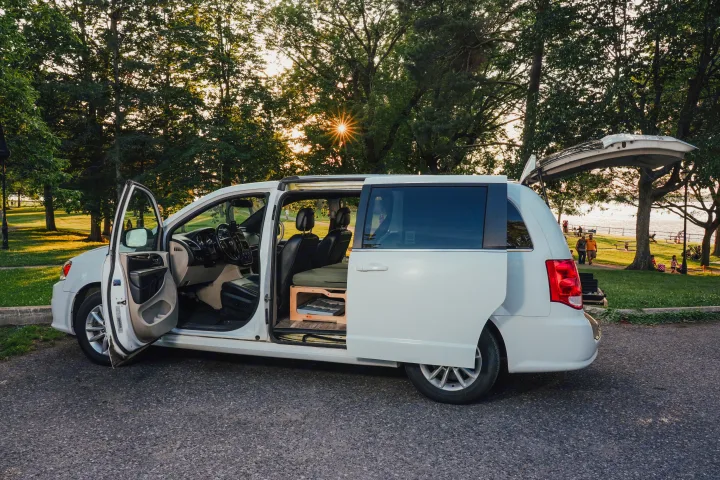 White minivan camper for RV rental, modified for travel, parked by a sunny park with people and water. Doors open.. Dodge Grand Caravan 2019