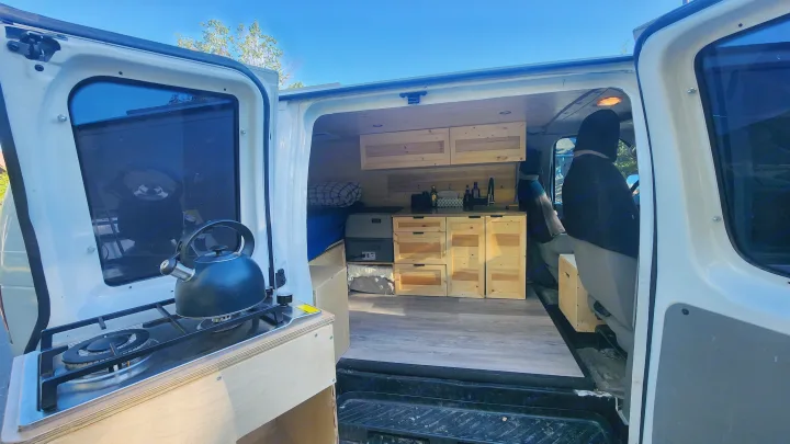 Inside a camper van, an open side door reveals a custom wood interior with a kitchenette and stove, ready for RV rental travel.. Ford Econoline 2011