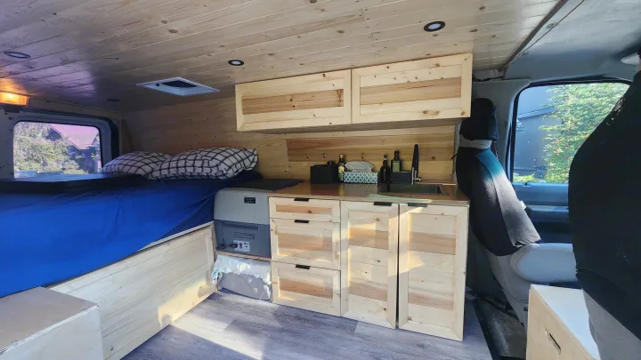 Cozy wood-paneled rv rental interior, featuring a bed with blue sheets, a kitchenette with sink, and storage cabinets.. Ford Econoline 2011