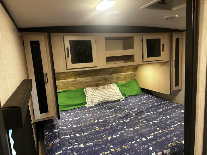 Cozy RV rental bedroom with a bed, camper-themed comforter, overhead cabinets, and wood-paneled headboard.. Heartland RVs Mallard 2022