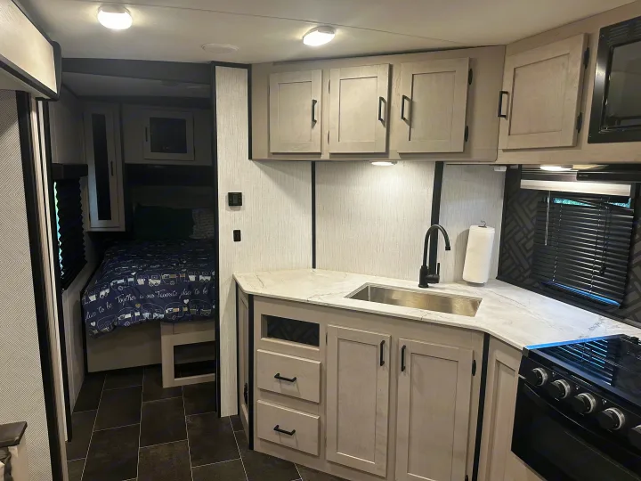 Bright RV rental interior showing a compact kitchen with sink & stove, and a comfortable bedroom area for travel.. Heartland RVs Mallard 2022