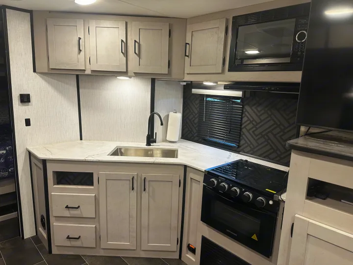 Modern RV rental kitchen with light gray cabinets, white marble-look counter, black sink, stove, microwave, and TV.. Heartland RVs Mallard 2022