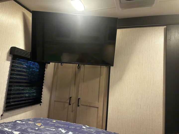 Inside an RV rental, a mounted TV overlooks a closet & bed. A dark window blind is on the left wall of this compact space.. Heartland RVs Mallard 2022
