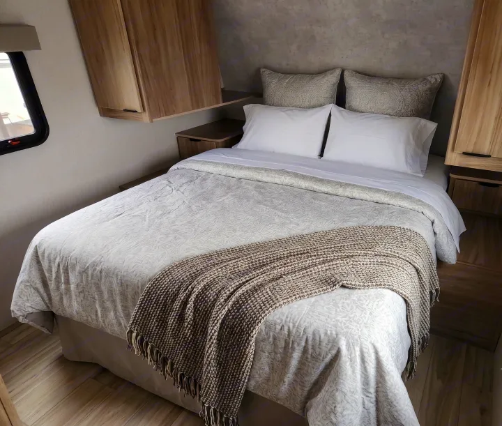RV rental bedroom featuring a comfy bed with white pillows, a patterned duvet, and a textured throw blanket, flanked by wood cabinets.. Winnebago Access 2025