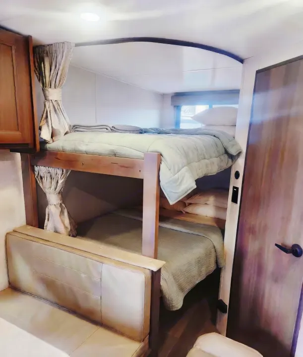 An RV rental's cozy bunk beds, complete with light blue comforters, tied-back curtains, and a nearby seating area.. Grand Design Transcend Xplor 2025