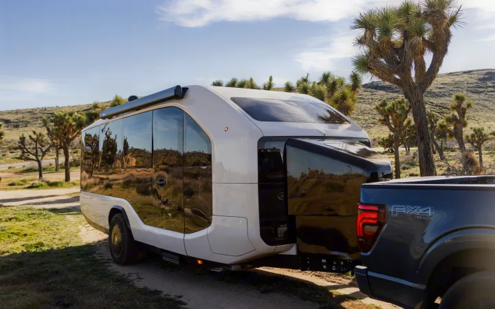 A modern white rv rental trailer is hitched to a gray pickup truck, parked among Joshua trees in a sunny desert.. Pebble Flow 2026