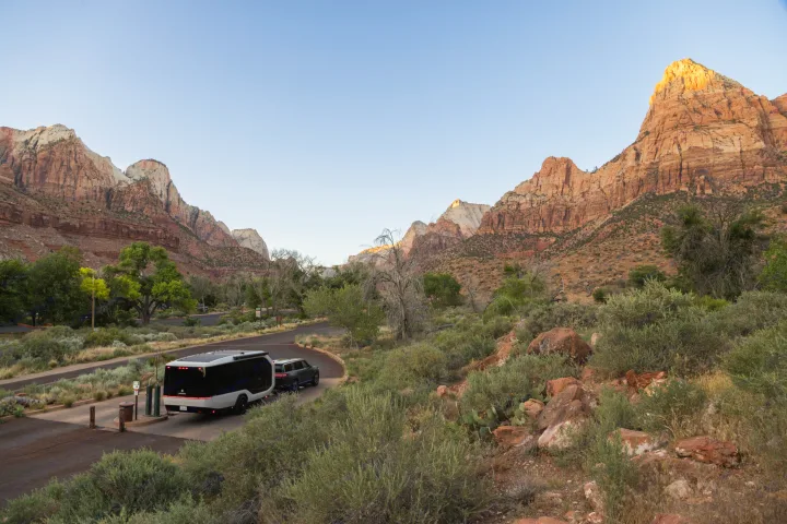 RV rental in Zion National Park: A sleek RV and SUV at a utility hookup, with sunlit red rock mountains and blue sky.. Pebble Flow 2026