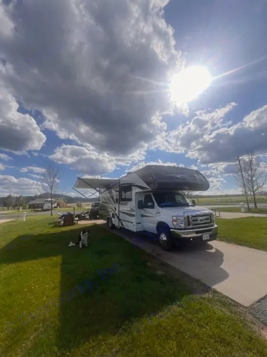 Enjoying a sunny day with an RV rental at a scenic campground. Awning extended, dog relaxing on the green grass.. Gulf Stream Conquest 2023