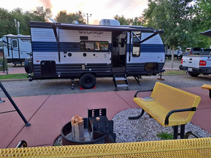A cozy RV rental setup with a Wolf Pup travel trailer, open awning, fire pit, and a yellow bench with books.. Forest River Cherokee Wolf Pup 2023