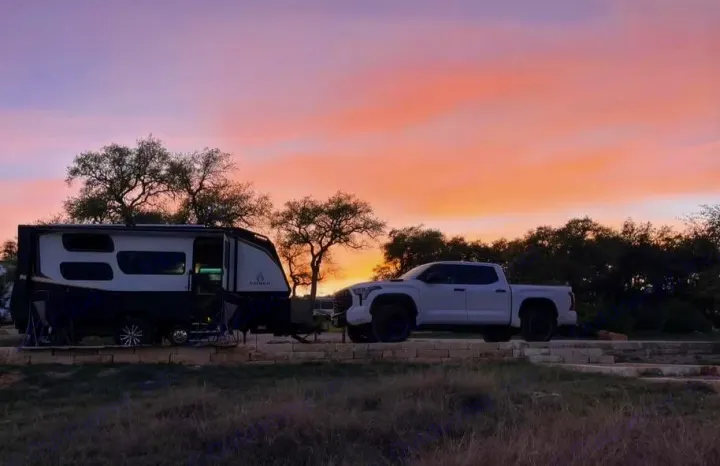 Our RV rental, a modern travel trailer, hitched to a white pickup at a beautiful sunset campsite, ready for adventure.. Ember Overland 2023