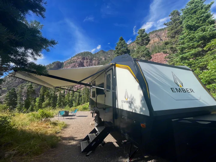 Enjoy a scenic Ember RV rental adventure! This travel trailer with an extended awning is set up at a mountain campsite, complete with chairs and a fire pit under a blue sky.. Ember Overland 2023