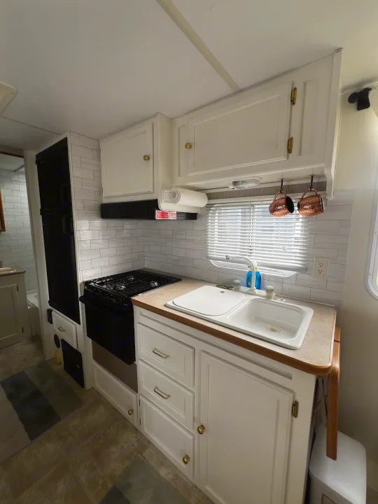 Cozy RV rental kitchen: white cabinets, subway tile, sink, stove, fridge, copper mugs, & bright window.. Kustom Koach Westwind 2004