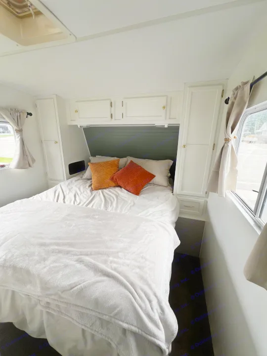 Bright RV rental bedroom interior with a neatly made bed, orange pillows, white cabinets, and windows.. Kustom Koach Westwind 2004