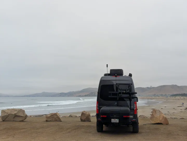 Black RV rental at an overcast beach, overlooking ocean waves, surfers, and distant hills. Features a bike rack and storage box.. Thor Sanctuary 2022