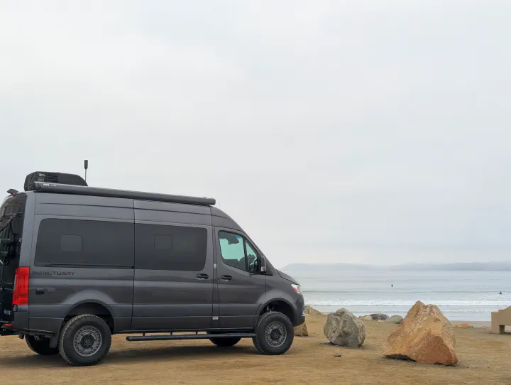 Grey Sanctuary RV rental on a sandy beach. Ocean waves, surfers, and rocks under a cloudy sky.. Thor Sanctuary 2022