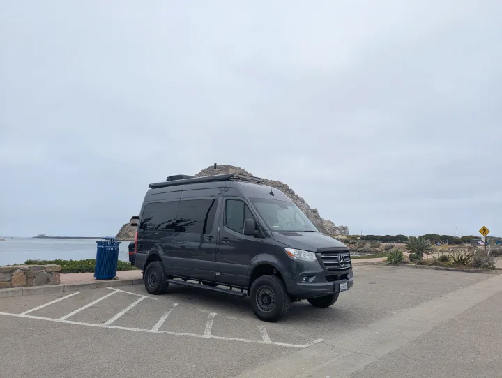 An all-terrain dark grey Mercedes-Benz Sprinter camper van, great for rv rental, parked in a coastal lot under a grey sky.. Thor Sanctuary 2022