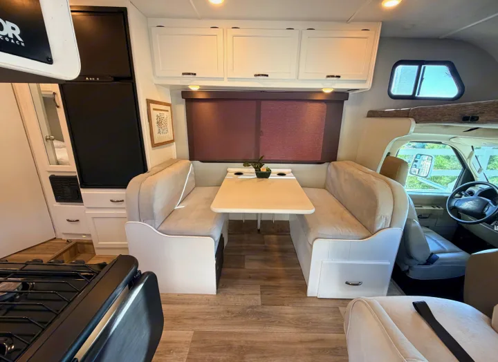 This rv rental interior features a cozy dinette, kitchen area with a fridge and stove, and overhead cabinets. The driver's cabin is visible upfront.. Thor Four Winds 2019