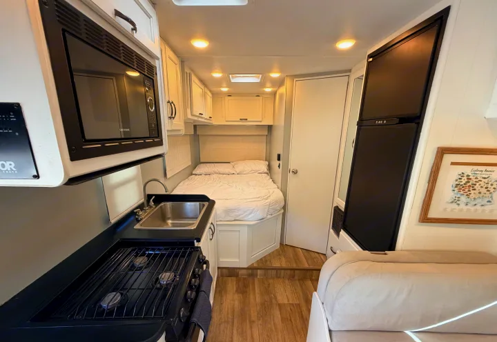 This bright RV rental interior features a compact kitchen with microwave, sink, stove, a comfy bed, & fridge.. Thor Four Winds 2019