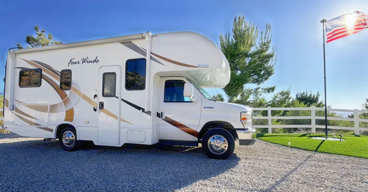 A white Four Winds motorhome on a sunny day. This RV rental is ready for adventure, parked on gravel with an American flag nearby.. Thor Four Winds 2019
