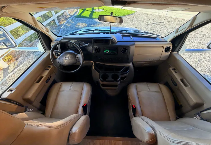 Inside an RV rental: wide view of the tan front cabin, showing the steering wheel, dashboard, seats, and windshield.. Thor Four Winds 2019