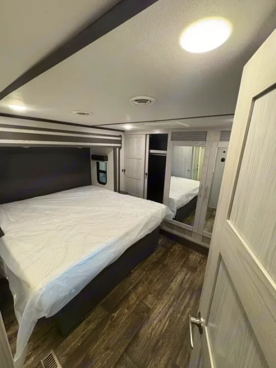 Modern RV rental bedroom with a queen bed (plastic wrapped), wood-look floor, and mirrored closet. Ideal for your journey.. Keystone Fuzion 2021