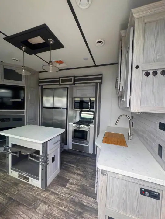 Modern RV rental kitchen featuring light wood cabinets, stainless steel appliances, a center island with seating, and spacious countertops.. Keystone Fuzion 2021