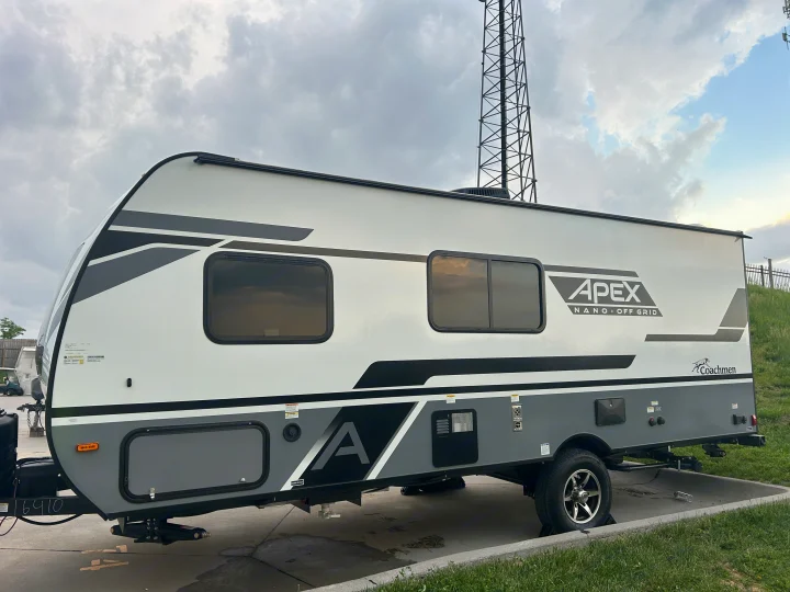 This white & gray Apex Nano off-grid travel trailer, an RV rental, is parked outdoors under a cloudy sky near a cell tower.. Coachmen Apex Nano 2024