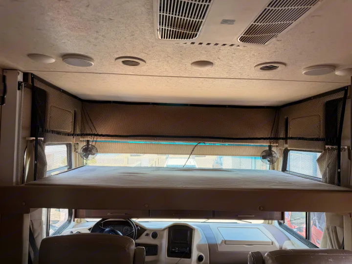 RV rental interior: large over-cab bunk bed with safety net and fans, dashboard, ceiling AC vents, and speakers.. Coachmen Mirada 32SS 2019