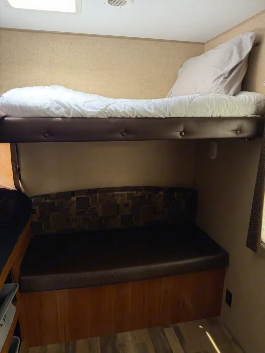Inside an RV rental: a compact sleeping area with an upper bunk bed and a lower cushioned bench, set against textured walls.. Starcraft Autumn Ridge 2015