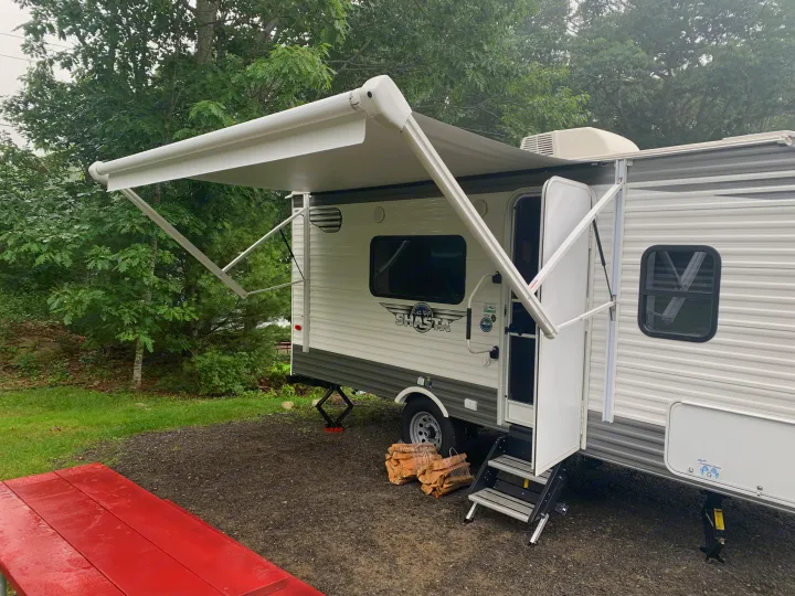 White and grey rv rental travel trailer with an extended awning at a gravel campsite, a red picnic table is in the foreground.. Forest River Shasta 2021