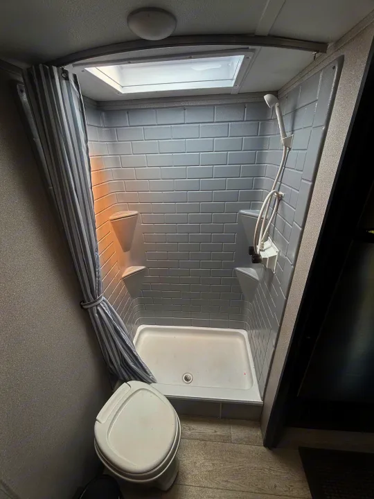 RV rental compact bathroom featuring a shower stall with subway-style tiled walls, corner shelves, and a toilet. A skylight brightens the space.. Forest River Shasta 2021