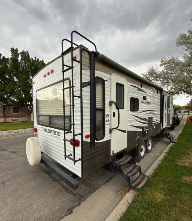 A white Wildwood travel trailer, with a rear ladder and spare tire, parked on a street. Ideal for your next RV rental adventure.. Forest River Wildwood 2017