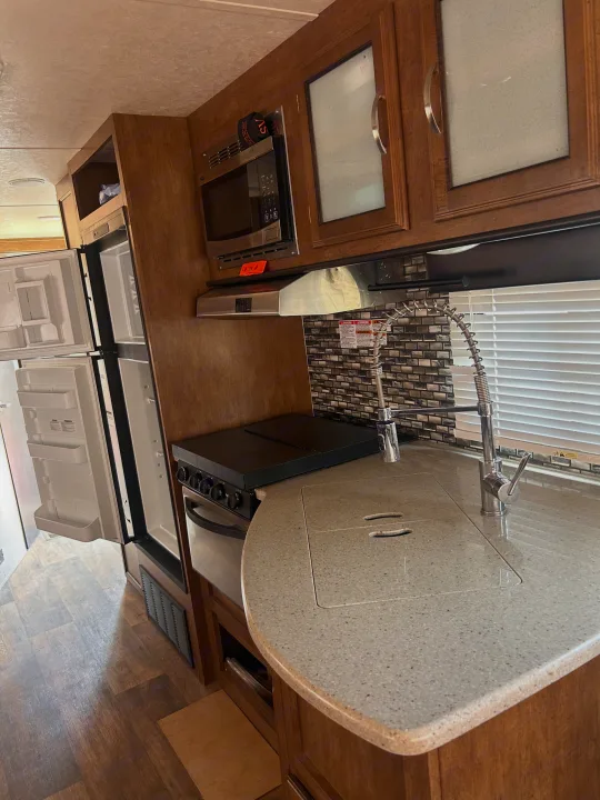 Inside a clean rv rental kitchen: open fridge, microwave, stove, sink, wood cabinets, and gray tiled backsplash. Modern & equipped.. Forest River Wildwood 2017