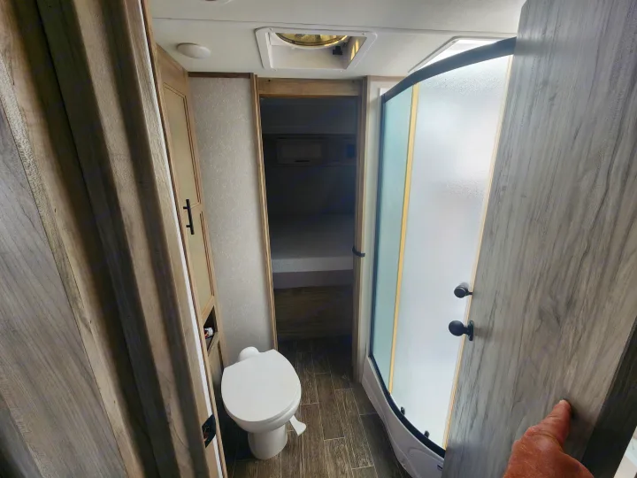 Inside an RV rental: a compact bathroom features a white toilet, curved shower, and open doorway leading to a bedroom area.. Forest River Cherokee Alpha Wolf 2021