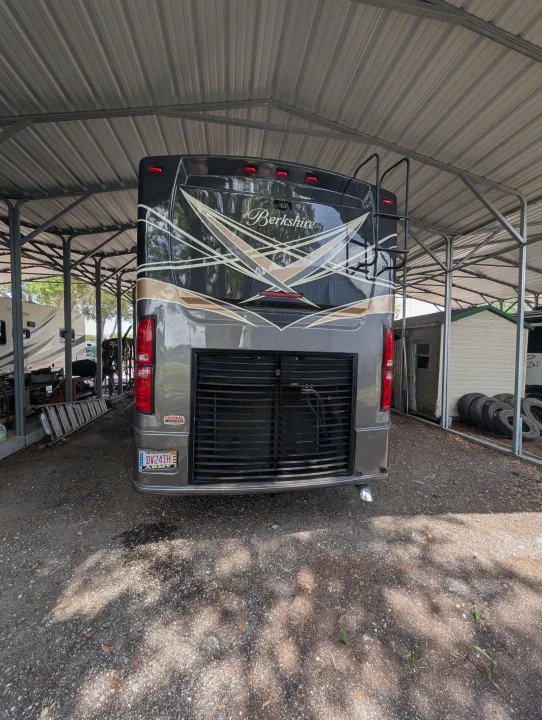 Rear view of a Berkshire RV under a metal carport, ideal for rv rental or storage. Gravel lot, US Army plate visible.. Forest River Berkshire 2018