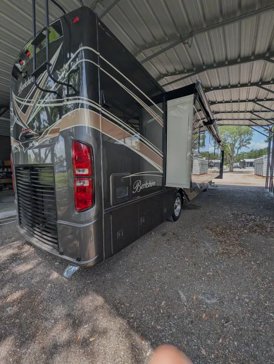 A dark Berkshire RV rental with its slide-out extended is parked on gravel under a metal shelter, ready for road trips.. Forest River Berkshire 2018