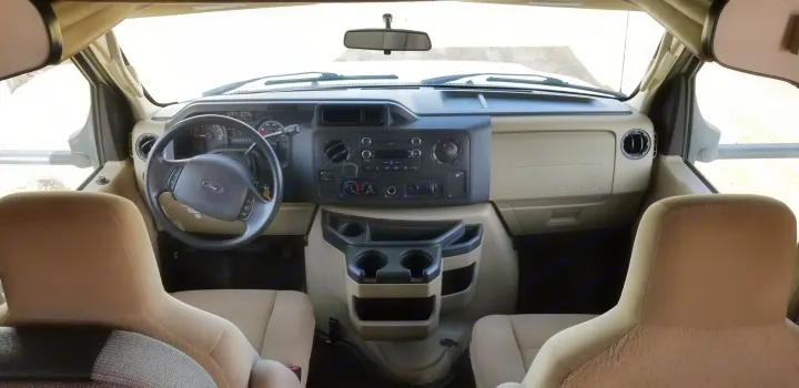 Inside an RV rental, showing the driver's cockpit, dashboard, and steering wheel from behind the beige seats. A clear view out the windshield.. Thor Majestic 2013