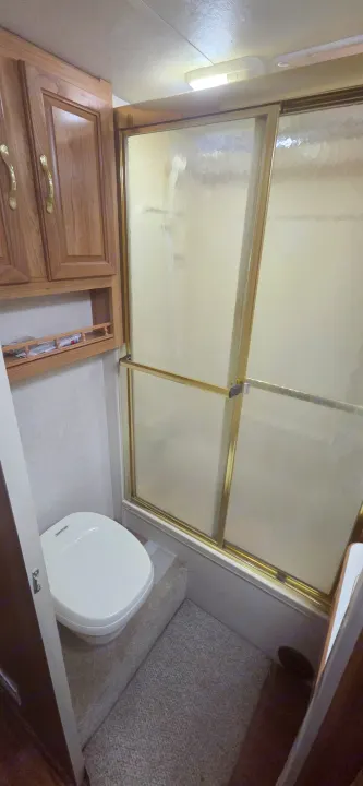 Compact RV rental bathroom showing a white toilet on a platform, frosted shower with gold frame, wood cabinets, and carpet.. Coachmen Leprechaun 2009