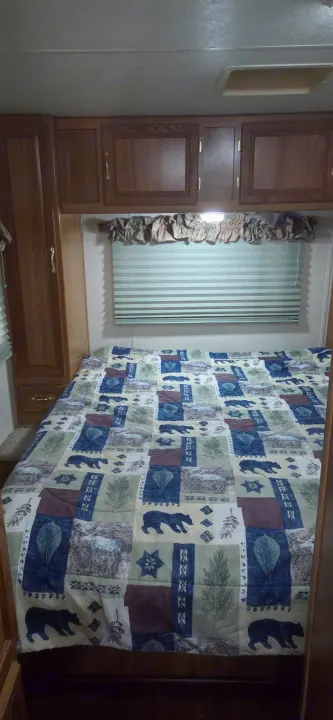 Cozy rv rental bedroom features a nature-themed comforter, wooden cabinets, and a window with blinds.. Coachmen Leprechaun 2009