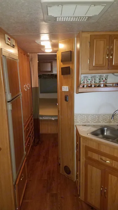Interior of an RV rental showing a galley kitchen with wood cabinets, sink, and refrigerator, and a hallway leading to a bedroom.. Coachmen Leprechaun 2009