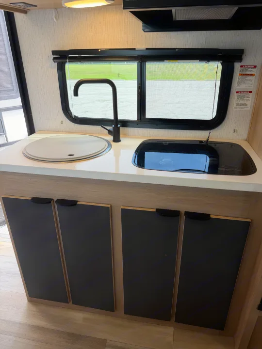 Modern RV rental kitchen with a covered sink, black faucet, glass-top stovetop, window view, and lower cabinets.. Cruiser RV Hitch 2022
