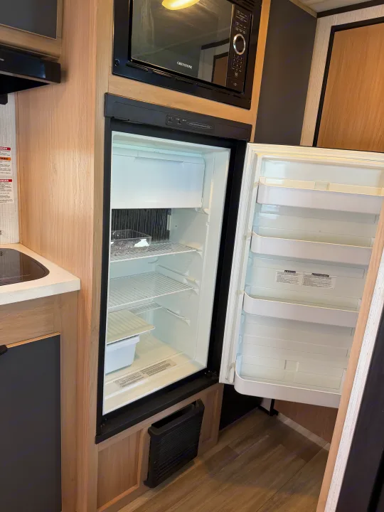 RV rental kitchen: An open, clean refrigerator with wire shelves and door bins, plus a microwave, set in light wood cabinetry.. Cruiser RV Hitch 2022