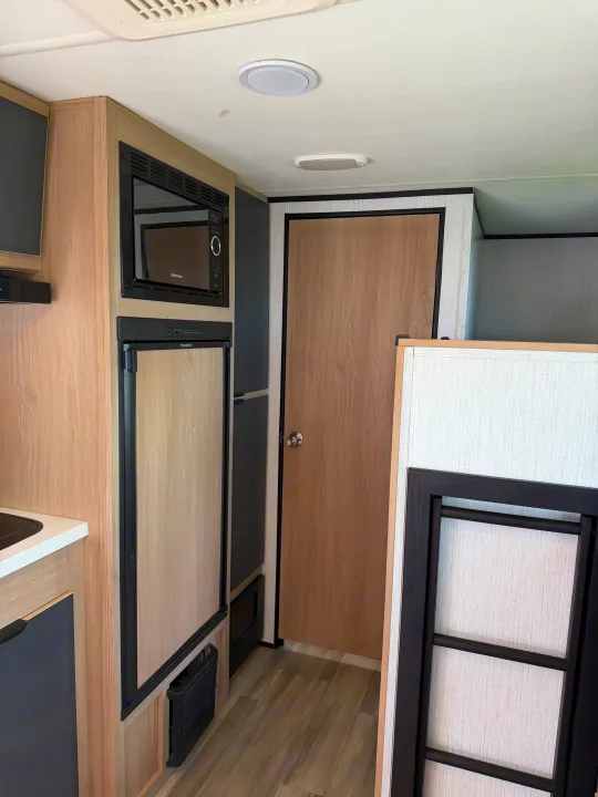 RV rental interior: A compact kitchen with a microwave, Dometic fridge, wood cabinets, and part of a bunk bed ladder.. Cruiser RV Hitch 2022