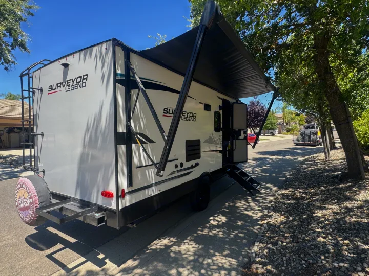 A white Surveyor Legend travel trailer, perfect for rv rental adventures, parked with its awning extended on a sunny day.. Forest River Surveyor Legend 2023