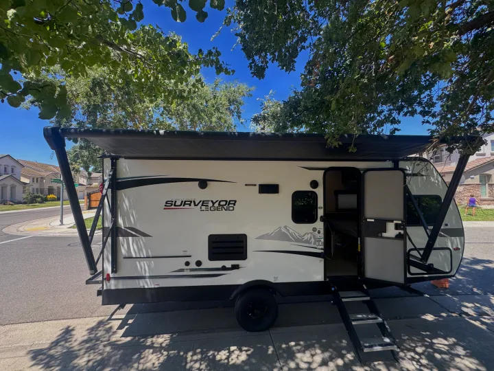 Surveyor Legend travel trailer RV rental parked under trees with awning extended and door open on a residential street.. Forest River Surveyor Legend 2023