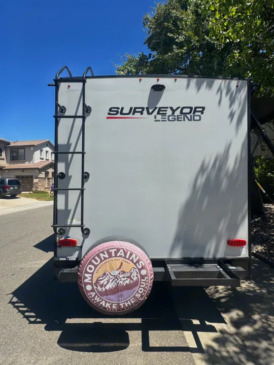 Rear of a Surveyor Legend rv rental travel trailer, with a ladder, mountain-themed spare tire cover, and cargo rack.. Forest River Surveyor Legend 2023