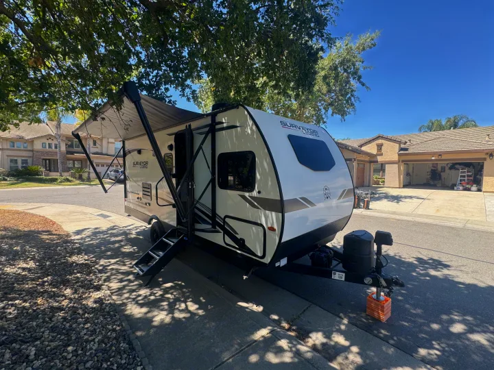 This white & black travel trailer rv rental is parked with its awning extended and steps down on a sunny residential street.. Forest River Surveyor Legend 2023